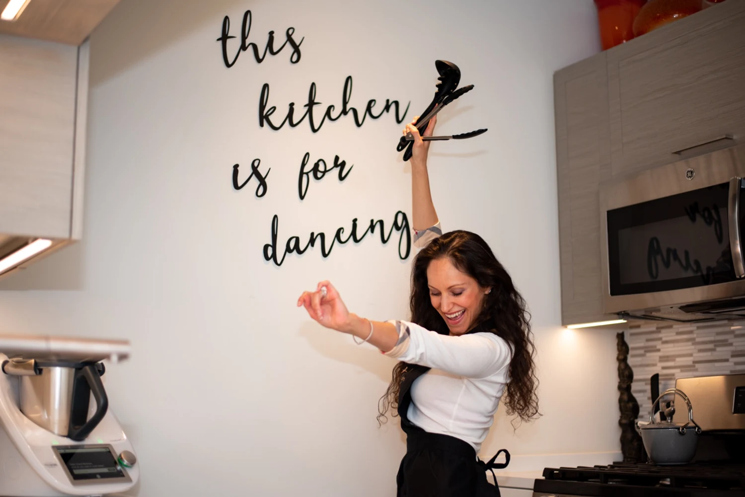 Cheryl dancing with cooking utensils beneath the words this kitchen is for dancing
